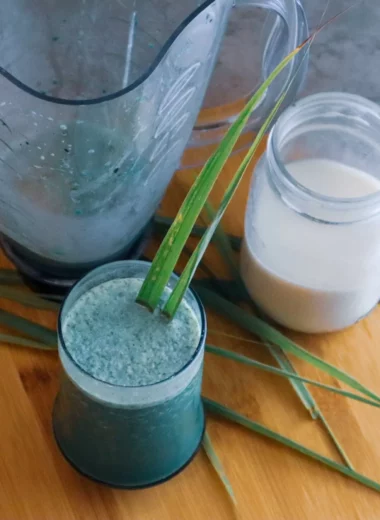 Fever grass oat milk smoothie in a glass with a blender and oat milk in the background.