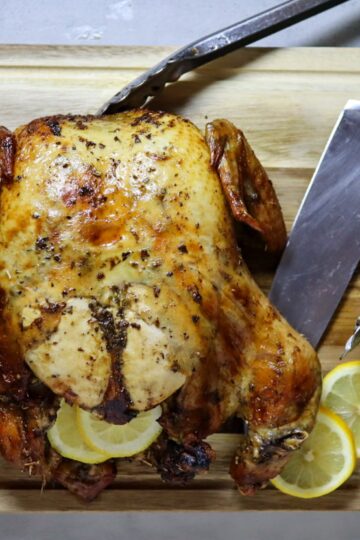 Lemon herb air fryer whole roasted chicken on a cutting board with fresh lemon slices.
