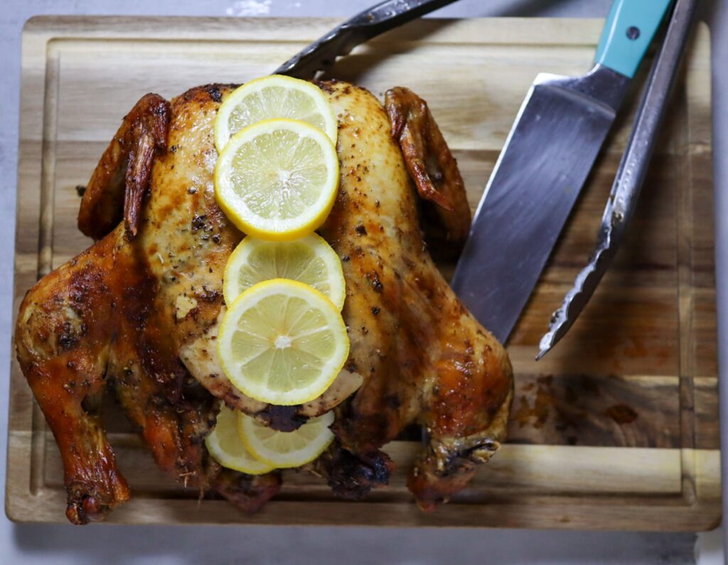 Lemon herb air fryer whole roasted chicken on a cutting board with fresh lemon slices.